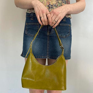 Olive green faux leather shoulder bag displayed with denim mini skirt outfit, featuring a sleek Y2K-inspired hobo silhouette and adjustable strap. Trendy minimalist handbag perfect for everyday wear, casual street style, vintage-inspired fashion, clean girl aesthetic, it-girl outfits, and modern women’s accessories. Chic olive purse with spacious interior and timeless design.