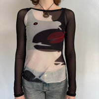 Front view of black sheer mesh graphic top for women with long sleeves, fitted stretchy Y2K going out top, abstract face print cool girl blouse, trendy boutique fashion shirt paired with denim jeans, sheer streetwear top.