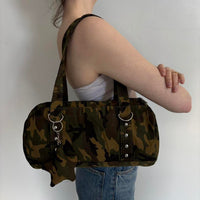 Side view of a Y2K camouflage shoulder bag featuring silver hardware, ring accents, and charm details. Vintage-inspired camo canvas purse with a structured silhouette and spacious interior, styled with a white tube top and denim jeans. Trendy army green shoulder handbag perfect for streetwear fashion, grunge outfits, Pinterest-inspired style, casual everyday wear, concerts, travel, and downtown It Girl aesthetics.
