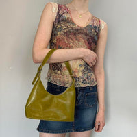 Model styling an olive green faux leather shoulder bag with adjustable strap, paired with a denim mini skirt and vintage-inspired mesh top. Trendy Y2K handbag featuring a minimalist hobo silhouette, perfect for everyday wear, casual outfits, indie sleaze fashion, clean girl aesthetic, streetwear looks, and modern it-girl style. Olive green purse with a sleek design and timeless fashion appeal.