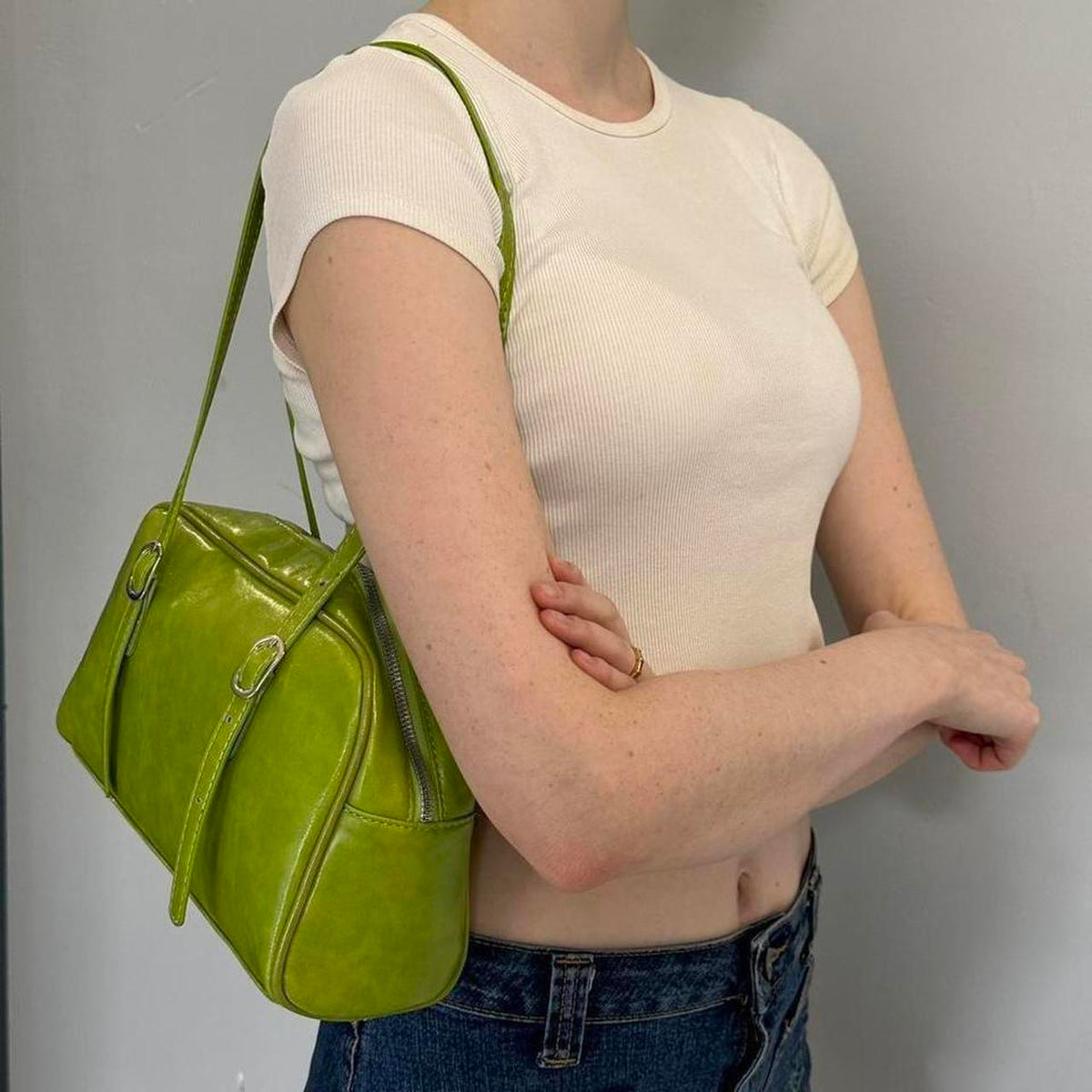 Lime green Y2K shoulder bag worn on shoulder, glossy faux leather baguette purse with adjustable buckle straps, vintage-inspired 2000s mini handbag styled with white ribbed crop top and denim jeans, trendy retro aesthetic accessory, statement street style bag for women