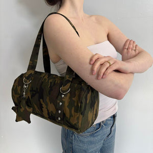 Y2K camouflage shoulder bag with silver hardware and charm detail, styled with a white tube top and denim jeans. Vintage-inspired camo purse featuring a spacious canvas design, trendy streetwear aesthetic, and army green camouflage print. Perfect everyday handbag for Y2K fashion, grunge outfits, Pinterest