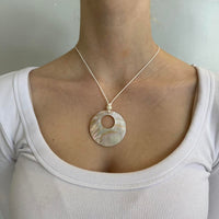 Close-up of mother of pearl shell pendant necklace with iridescent circular charm on a minimalist cord chain, styled with a white ribbed scoop neck top. Coastal cool girl jewelry inspired by clean girl fashion, Y2K aesthetics, beachy outfits, and Pinterest-style minimalist accessories.