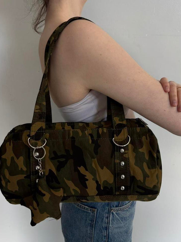 Side view of a Y2K camouflage shoulder bag featuring silver hardware, ring accents, and charm details. Vintage-inspired camo canvas purse with a structured silhouette and spacious interior, styled with a white tube top and denim jeans. Trendy army green shoulder handbag perfect for streetwear fashion, grunge outfits, Pinterest-inspired style, casual everyday wear, concerts, travel, and downtown It Girl aesthetics.